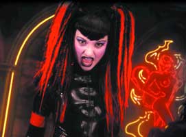 A person with long red and black hair and dark makeup poses in front of a neon sign.