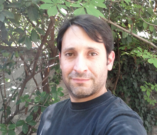 A man with dark hair and light stubble wearing a black t-shirt, positioned in front of green leafy plants.