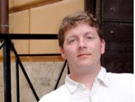 A man with light brown hair wearing a white collared shirt looking forward.