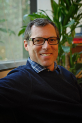 A smiling man with short dark hair and glasses, wearing a navy sweater over a blue checkered shirt.
