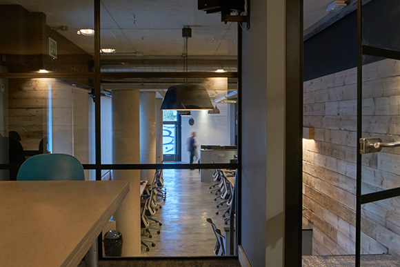 View through a glass wall into a brightly lit office with desks, chairs, and large concrete pillars.