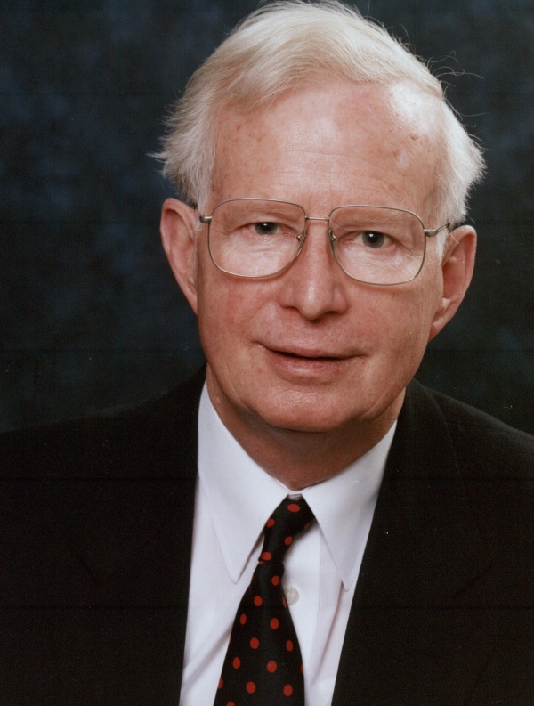 An older man with white hair and glasses wearing a dark suit, white shirt, and red polka dot tie.