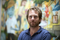 A man with short brown hair and a beard wearing a blue polo shirt, standing before a vibrant mural.