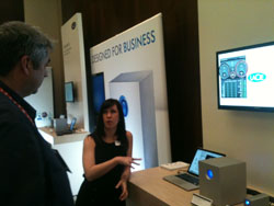A woman in a black dress gestures while presenting a product at a trade show booth. A man stands to her left, facing her.