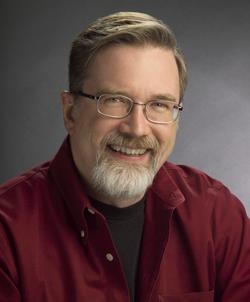 A man with glasses and a beard wearing a red shirt smiles against a dark background.