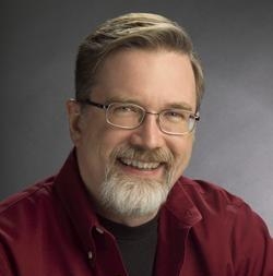A man with short brown hair, glasses, and a grey goatee smiling against a dark grey background.
