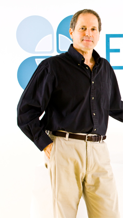 A man wearing a dark button-down shirt and khaki trousers stands against a white background with a blue logo.