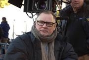 A man with glasses and a tan scarf stands outdoors in front of a large professional film camera.