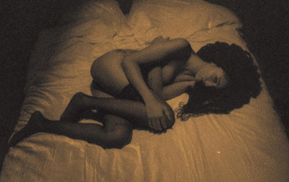 A person with long dark hair lies curled on a white bedsheet in dim, warm lighting.