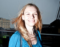 A woman with blonde hair and a blue shirt smiles at the camera. Her hair is blowing in the wind.