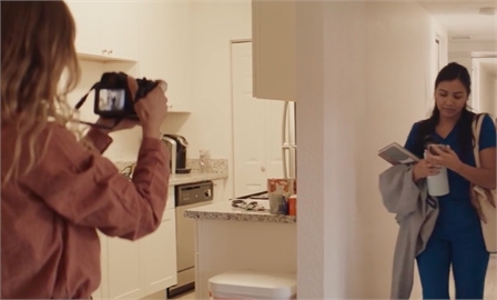 A person holds a camera to photograph a woman standing in a hallway holding a book and a mug.