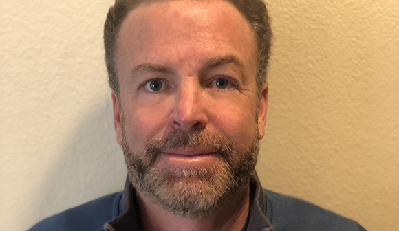 Close-up portrait of a man with light eyes and a salt-and-pepper beard looking directly at the camera.
