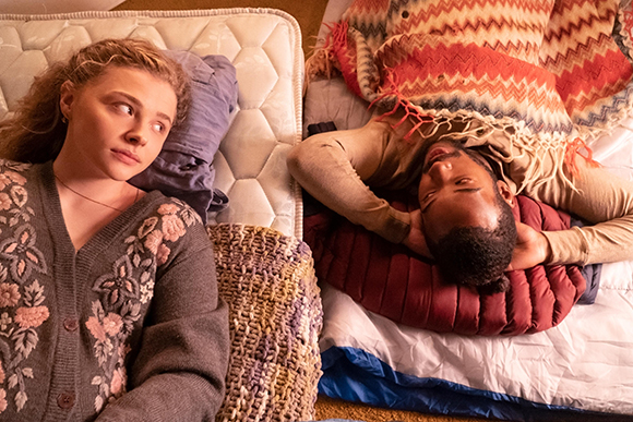 A blonde woman in a floral cardigan looks sideways while a man lies next to her under a patterned blanket.
