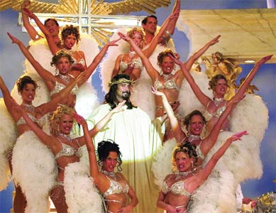 Performers in white feathered costumes and silver bikinis pose on a stage around a central figure.