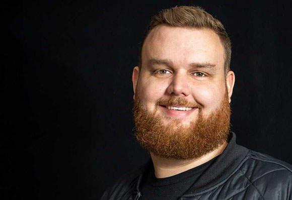 A smiling man with short brown hair and a full reddish-brown beard against a black background.