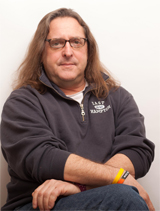 A man with long brown hair and glasses wearing a dark grey quarter-zip pullover.
