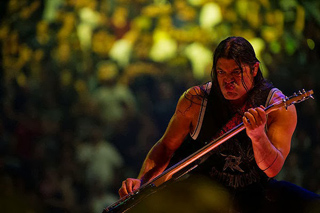 A man with long dark hair playing a bass guitar during a live performance under warm stage lighting.