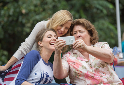 Three women laugh and smile while posing for a selfie with a smartphone in a park.