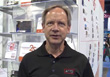 A man with light hair and blue eyes wearing a black polo shirt stands before a retail display.