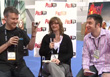 Three people sitting in chairs, holding microphones and a small camera, in front of a branded backdrop.