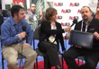 A woman holds a microphone toward a man who is holding a black tablet while seated.