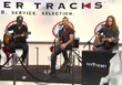 Three musicians perform on a stage in front of a white backdrop with text.