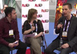 A man in a dark suit speaks while a woman holds a microphone and another man listens during an interview.