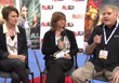 Three people sit in chairs on a stage in front of promotional posters and logos.