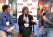 A man in blue, a woman in black, and a man in grey sit in chairs holding microphones.