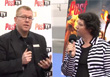 A man in a black suit and a woman in a striped jacket hold microphones during an interview.