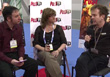 A man in a maroon shirt, a woman in a black top, and a man in a black shirt sit in a row talking.