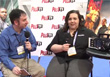 A man in a blue shirt holds a microphone while interviewing a woman in a black shirt.