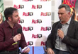 Two men sit facing each other, holding microphones while talking in front of a branded backdrop.