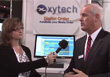 A woman holds a microphone toward a man in a suit standing in front of an xytech banner and a computer monitor.