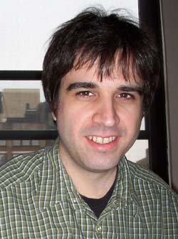 A man with dark hair and a green plaid shirt smiling in front of a window.