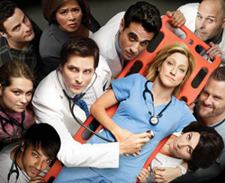 A group of doctors and nurses looking at the camera, surrounding a woman in blue scrubs on an orange stretcher.