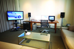 A living room featuring a desk with a computer, a large TV showing a car, and speakers on stands.