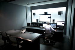 A person sits at a long desk with multiple computer monitors and speakers in a dimly lit control room.