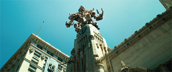 A large, complex metallic sculpture sits atop a tall, ornate stone building tower under a clear blue sky.