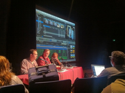 Three people sit at a table in a dark room facing a large projection of video editing software.