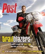 Man in motocross gear holding a dirt bike with a sunny sky in the background.