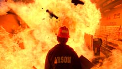 A person wearing a red hat and a shirt with 'ARSON' printed on the back faces a massive orange fireball.