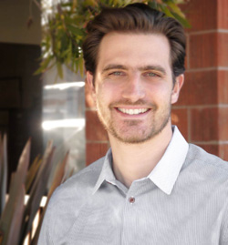 A smiling man with dark hair and light eyes wearing a grey collared shirt against a brick background.
