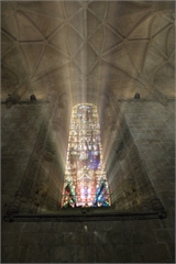 Sunlight beams through a tall, colorful stained glass window into a vaulted stone cathedral ceiling.