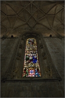 A tall, colorful stained glass window set within a dark stone wall under a complex ribbed vaulted ceiling.