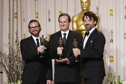 Three men in black formal attire smiling and holding gold Oscar awards in front of a large gold statue.
