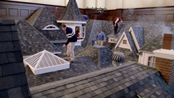 People walking across a large-scale miniature model of a grey shingled roof with dormers and chimneys.