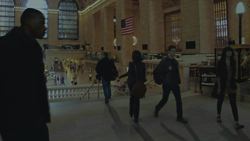 People walking through a large, dimly lit hall with high ceilings and an American flag hanging in the background.
