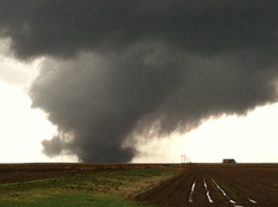 A dark tornado funnel descends from heavy storm clouds over a flat landscape.