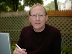 A man with glasses and a dark maroon shirt sits in front of a wooden lattice fence.